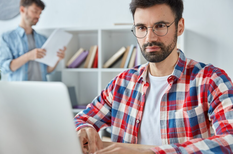 man in colorful shirt works on laptop, other man stands and reads the book behind him